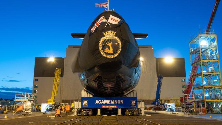 A black submarine with a banner below detailing 'AGAMEMNON'. The submarine stands outside a hangar at night with a bright blue sky.