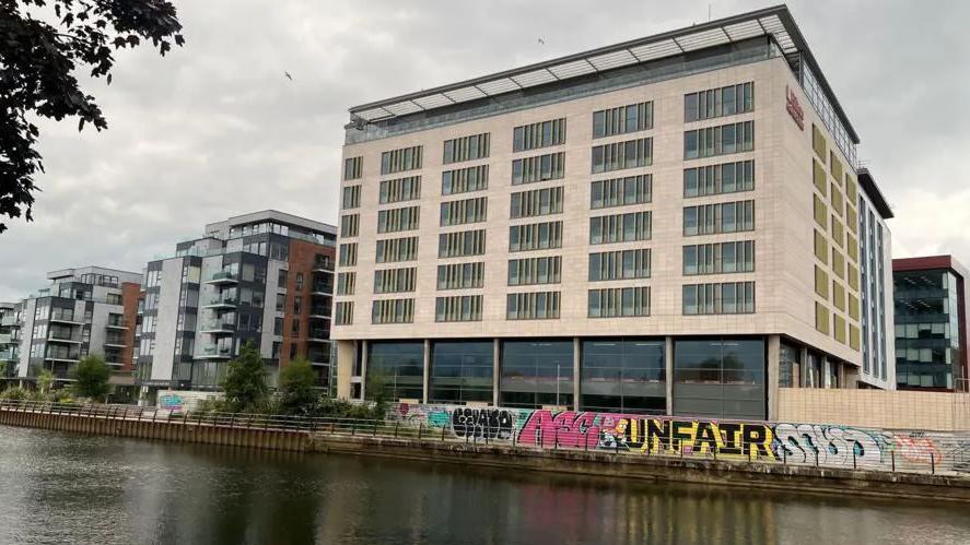 Exterior of the hotel building - a cream tall tower with glass windows - next to the River Nene with graffiti on boarding around the hotel.