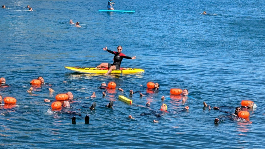 Students learn to float in the water while an instructor sits on a yellow surfboard. The instructor is wearing a wetsuit and has their arms outstretched.