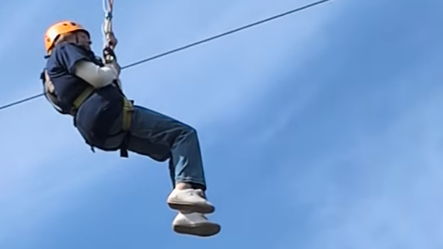 80-year-old Maureen Burton, wearing a T-shirt printed with her late husband's photograph and an orange safety helmet is hanging 13 metres in the air on a zip wire.
