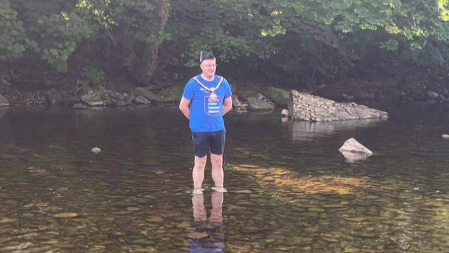 A man wearing a blue T-shirt, black shorts and mayoral chain and hat stands in a river.