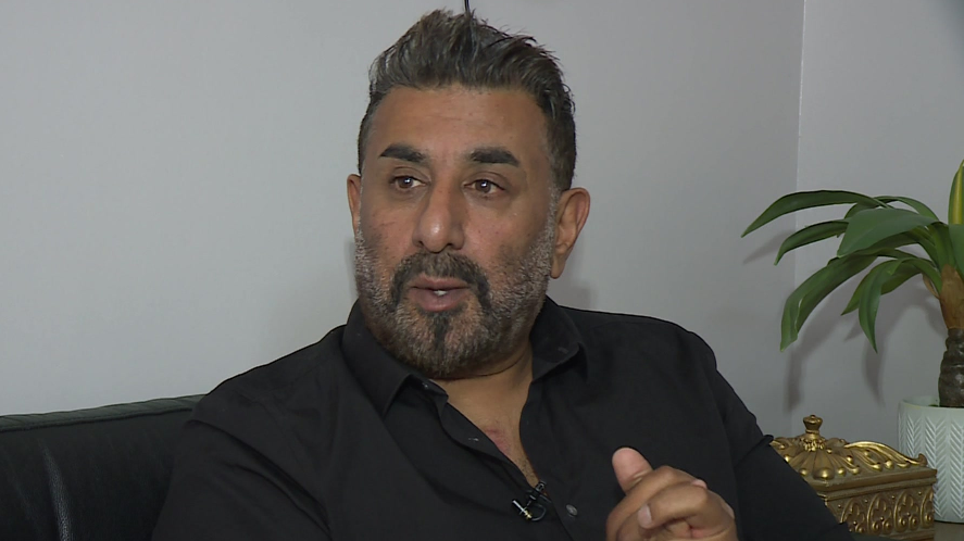 Shabaz Hussain sits on a sofa with his hands clasped. He wears a black shirt and has short black hair and a short black and grey beard. 