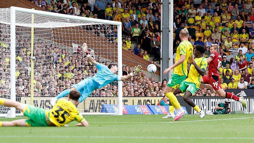 Aune Heggebo puts West Bromwich Albion in front against Norwich City. He is watching the ball as it evades the diving goalkeeper ready to put it in the net. Heggebo is all in red. There are two Norwich defenders in front of him wearing yellow shirts and green shorts. 
