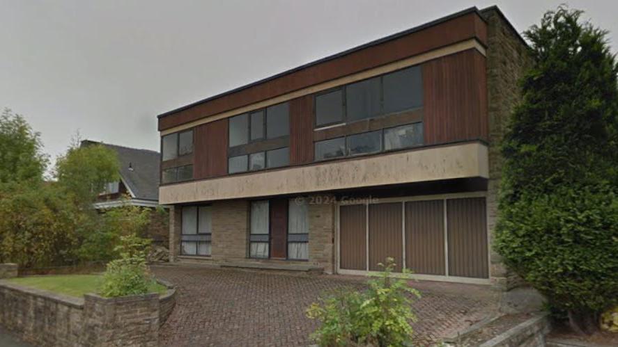 A large two-storey building with three windows and wooden cladding on the upper storey and two windows and garage doors on the ground floor. Block paving and a small grass area is in the foreground.
