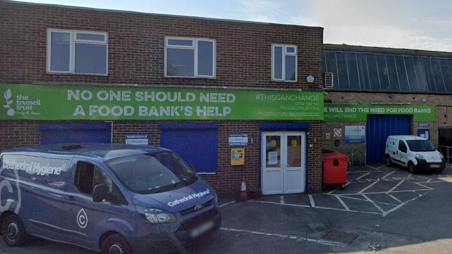 A view of a single-storey small warehouse in Salisbury, red brick. On it are big signs in green for the foodbank with the Trussell Trust logo. It says "No one should need a food bank's help".