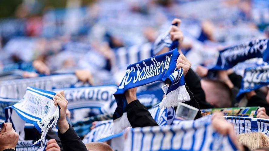 A number of fans holding blue and white FC Schalke 04 scarves at a game