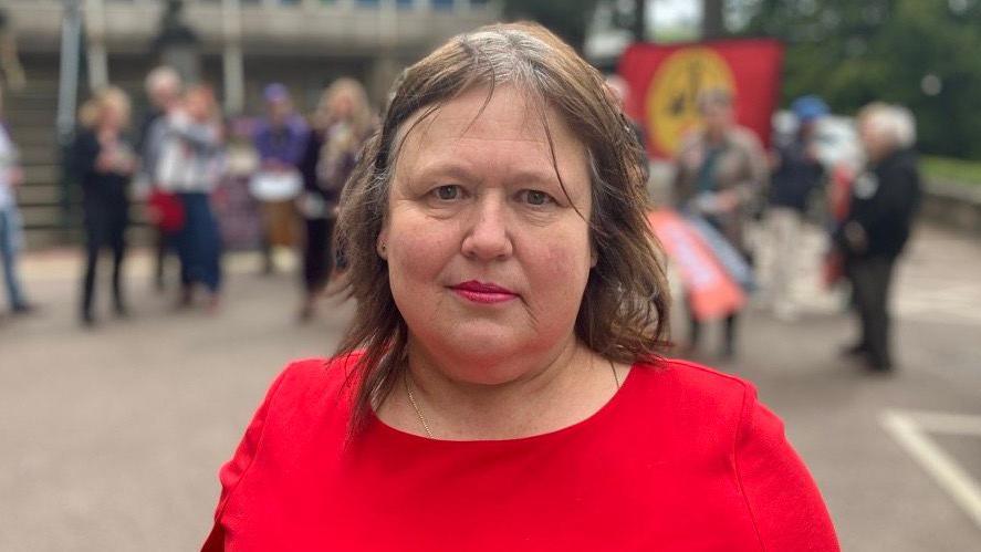 Head and shoulders shot of Joan Dixon - she wearing a red dress and is looking directly into the camera. Protestors outside County Hall in Matlock can be seen in the background