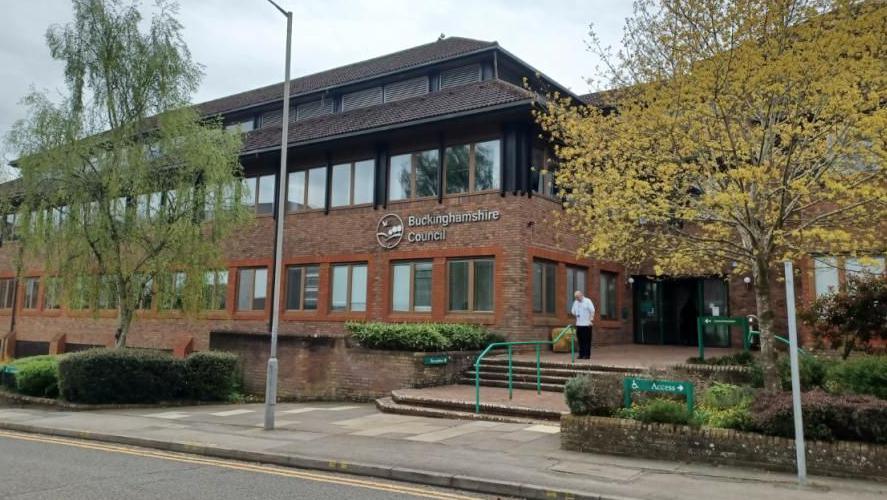 A brown three-storey building with "Buckinghamshire Council" in lettering on the front. The picture is taking from the other side of the road and shows steps leading up to the entrance, trees in front of the building, and a man about to walk down the steps.