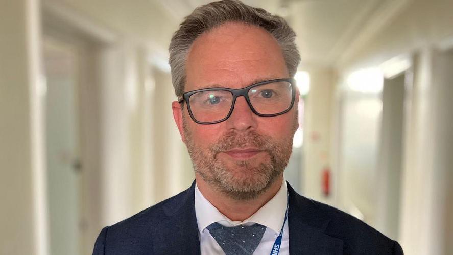 Head and shoulders picture of Martin Hodgson. He has greying hair, a short beard and glasses and is wearing a dark blue suit and tie with a white shirt and NHS lanyard