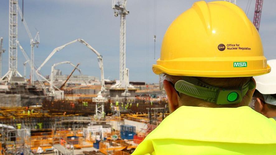 A person wearing a high-vis jacket and a yellow hard hat with the words 'Office for Nuclear Regulation' on the back looks over construction at the Hinkley C site. Lots of cranes and scaffolding can be seen covering the site, as well as a large number of workers in high-vis.