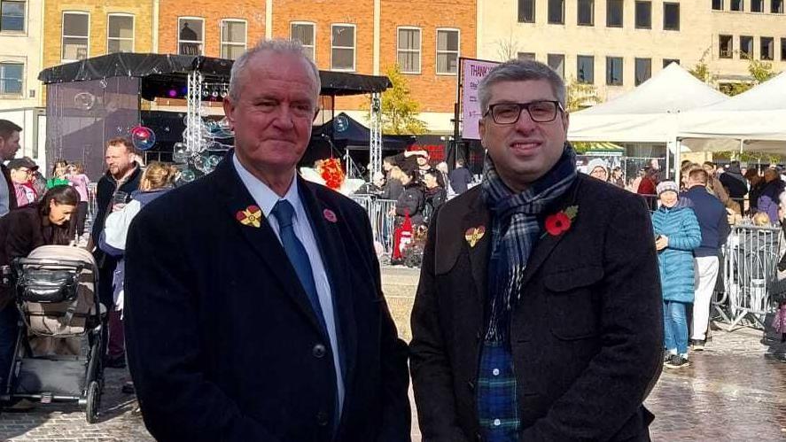 A man in a black wool coat, a white shirt and navy blue tie stands next to a man in a charcoal jakcet, and a navy tarten scarf. They are standing in front of several market stalls with lots of people waiting at them. An outdoor stage is set up behind them.