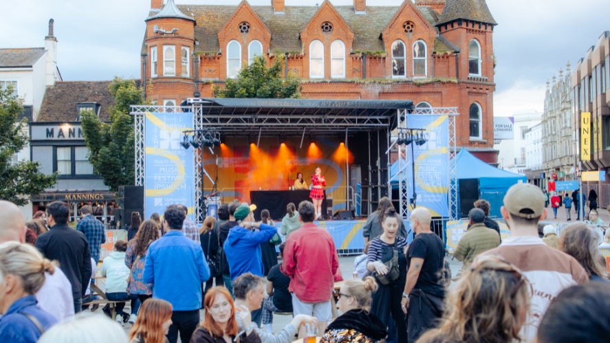 A pop-up stage is shown in the centre of the picture, with a singer in the middle of the stage wearing a red outfit. Many people are seen gathering to watch the performance.