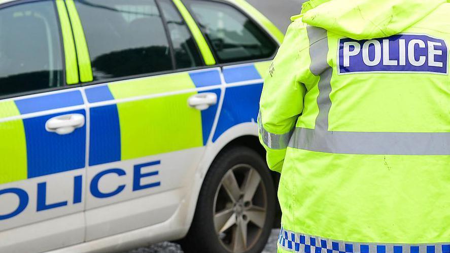 The picture shows the back of a police officer wearing a yellow luminous jacket and standing beside and yellow and blue coloured police car