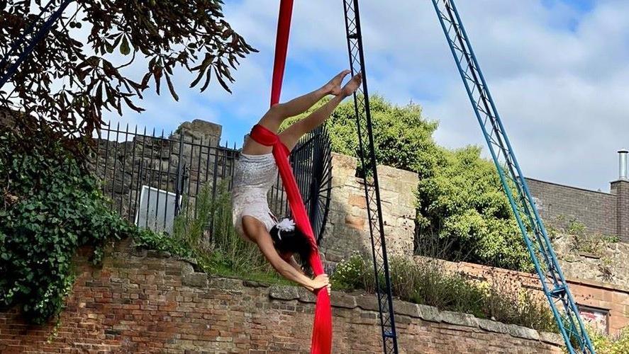 A trapeze artist upside down twirling on red material. Behind her is the wall of Tamworth castle and to the right is tall aerial equipment. 