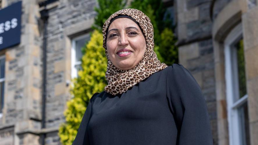 Nagina, who is wearing a leopard print headscarf, is smiling as she stands outside the Loch Ness Centre in sunshine.