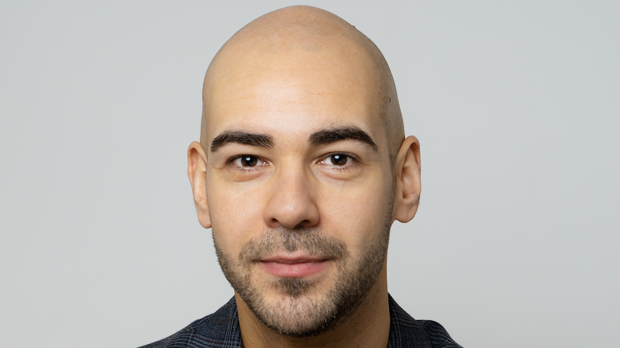 A bald man with stubble smiles without teeth at the camera wearing a black t-shirt and grey and white blazer. 