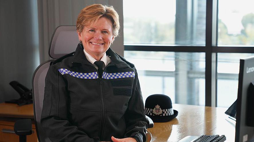 Maggie Blyth wearing a black police's uniform with blue and white checked stripe across the chest, with her police cap beside her. She is sitting at a desk in front of a computer with her elbows resting on the table. She has short blonde hair and is smiling at the camera.