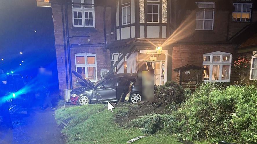 A smashed black BMW car with the hood popped open against a window and brick wall that is smashed.