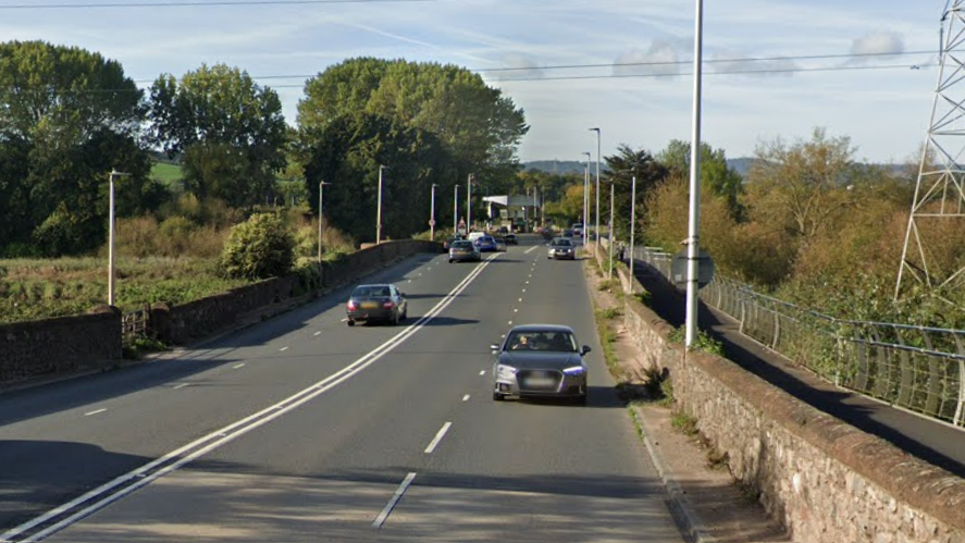 Google image of Bridge Road. Cars are travelling on either side of the carriageway. Trees and lampposts line each side of the road.