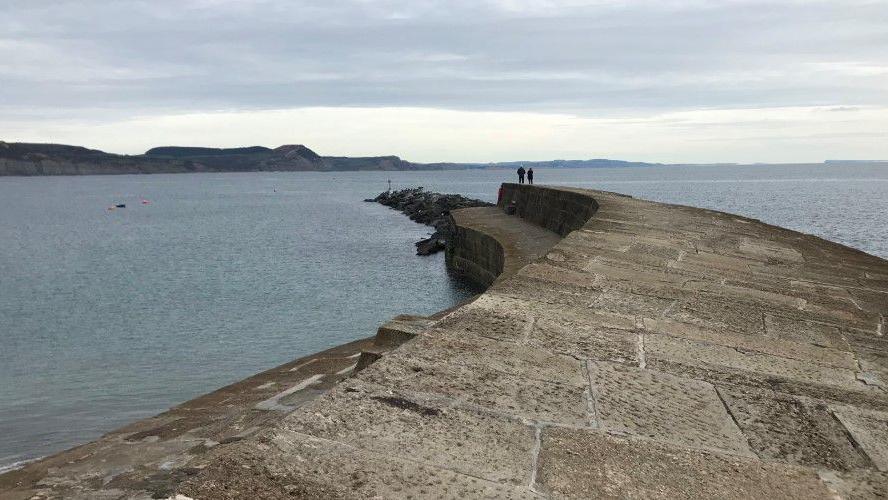 View from the Cobb looking east across the sea towards Portland. Two people are standing on the end of the wide, stone breakwater. In the distance are the cliffs of the Jurassic Coast.