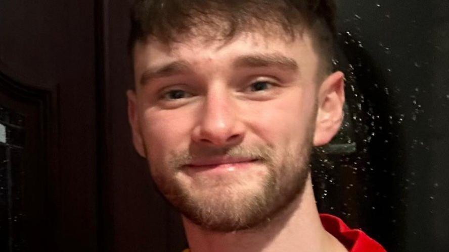 Kieran Thomas Hughes smiling into the camera. he has a short brown beard and brown hair with a fringe. He is wearing a red shirt.