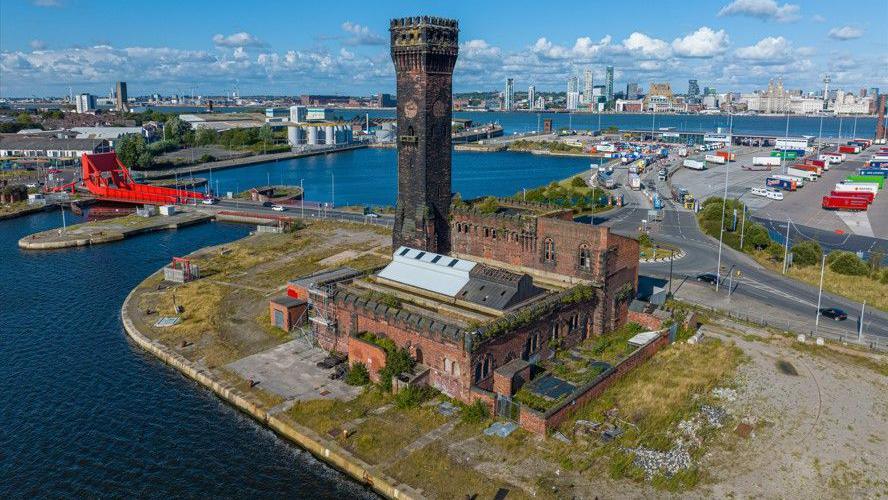 A photo of the Hydraulic Tower showing the squat, brick building from which the  tower itself rises. The tower is blackened with soot and the whole building has vegetation growing from it. It stands on an overgrown piece of dockland, and the Liverpool waterfront is visible across the Mersey in the background of the photo