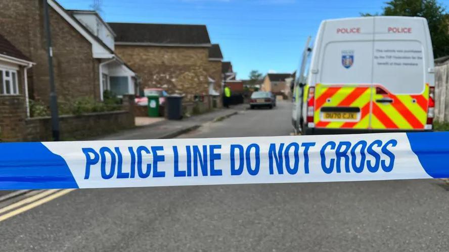 'Police line do not cross' tape across a street with a police van in the background