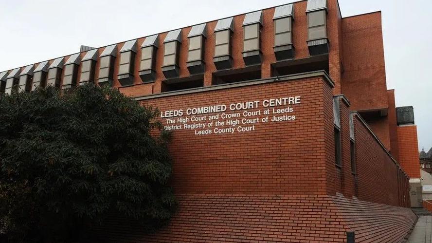 Leeds Crown Court - a redbrick building signposted as 'Leeds Combined Court Centre'. Underneath are the words: "The High Court and Crown Court at Leeds; District Registry of the High Court of Justice; Leeds County Court." A bush sits to the side.