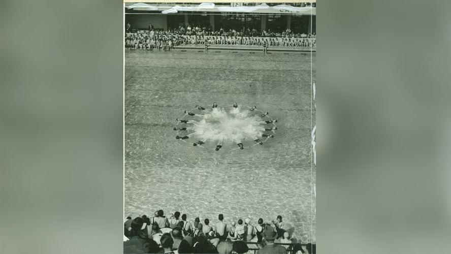 A black-and-white photo captures spectators watching a synchronised swimming display in Morecombe Swimming Stadium