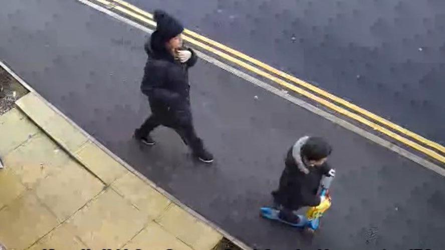 A CCTV still of Claire and Lincoln Button on a pavement outside their flat. They are wearing black coats. Lincoln is riding a yellow and blue scooter.