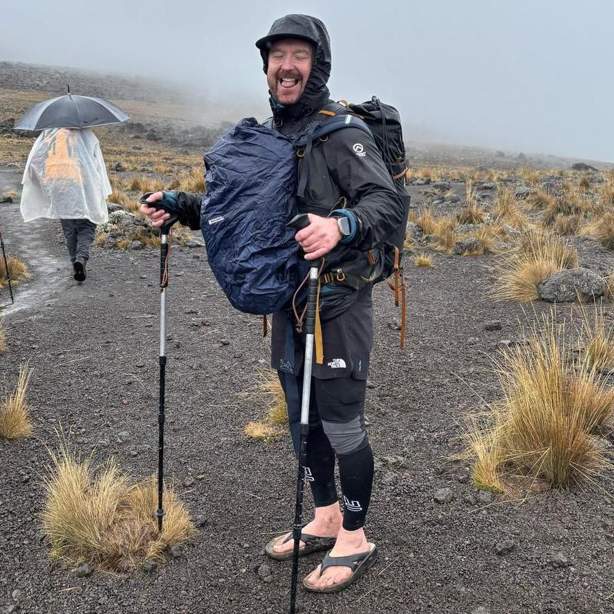 A laughing Stu Skinner pokes his tongue out for the camera during a rainy hike. He is wearing black hiking gear with a hood and carrying two full backpacks on his front and back. He is also holding Nordic hiking poles in his hands. His feet are bare with the exception of a pair of flip flops. The landscape is bare dark grey stone, dotted large spiky tufts of grass. A person in the background is wearing a poncho and carrying an umbrella.