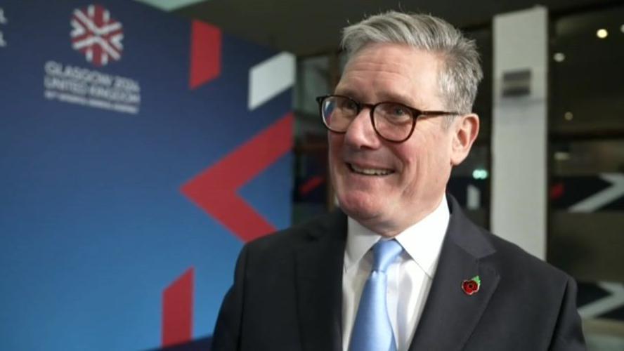 A screenshot of Keir Starmer talking during an interview. He is smiling, and wearing dark-framed glassed and a dark suit with a light blue tie. He is also wearing a poppy badge. 