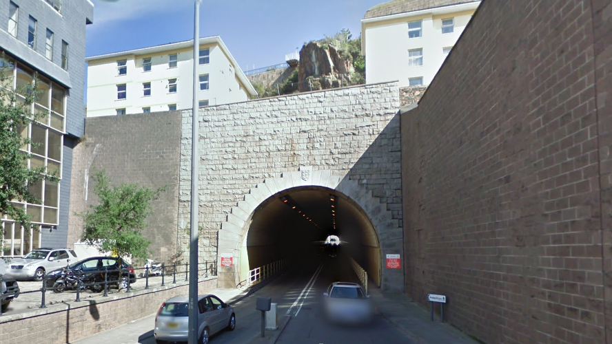 Fort Regent Tunnel on a sunny day with clear blue skies. One silver car is seen leaving the tunnel with a second silver car entering it. The entrance to the tunnel is made from grey stone.