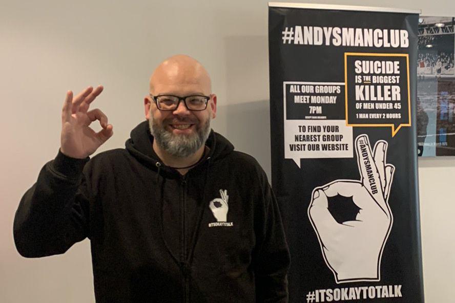 Simon Mayo, who is bald and wears glasses. He has a grey beard and is performing the OK symbol while stood in from of a banner promoting Andy's Man Club