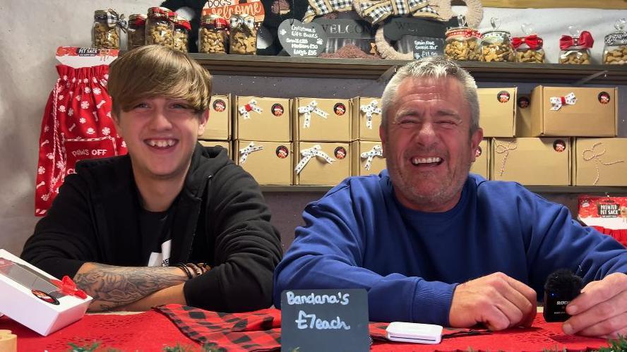 Two men standing and smiling side by side. The man on the left is younger, with long brown hair and a tattoo on his arm. The man on the right is laughing, wearing a blue sweatshirt and short grey hair. There are boxes piled behind them. 