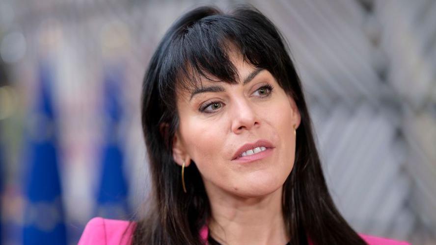 Jennifer Carroll MacNeill is staring off to the side. She has long black hair and is wearing gold earrings and wearing a pink suit. The background is blurred. 