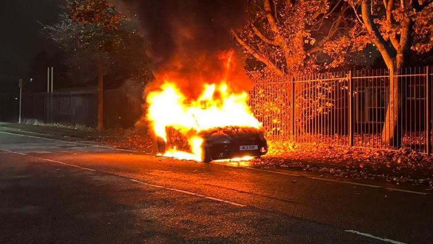 A car ablaze at night on the side of a street at night in Salford. Flames can be seen coming from the blackened vehicle, which is close to a fence and some trees.