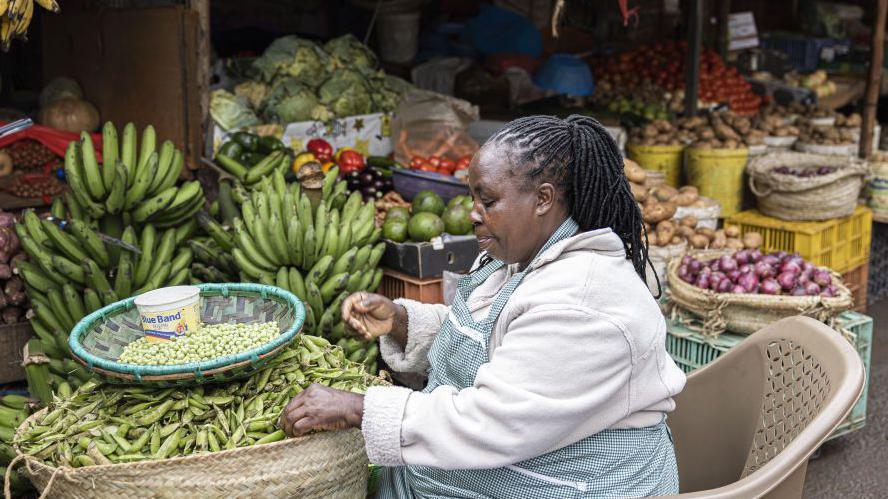 A woman, a vendor, at a market stall in Kenya with green peas, green bananas and other groceries