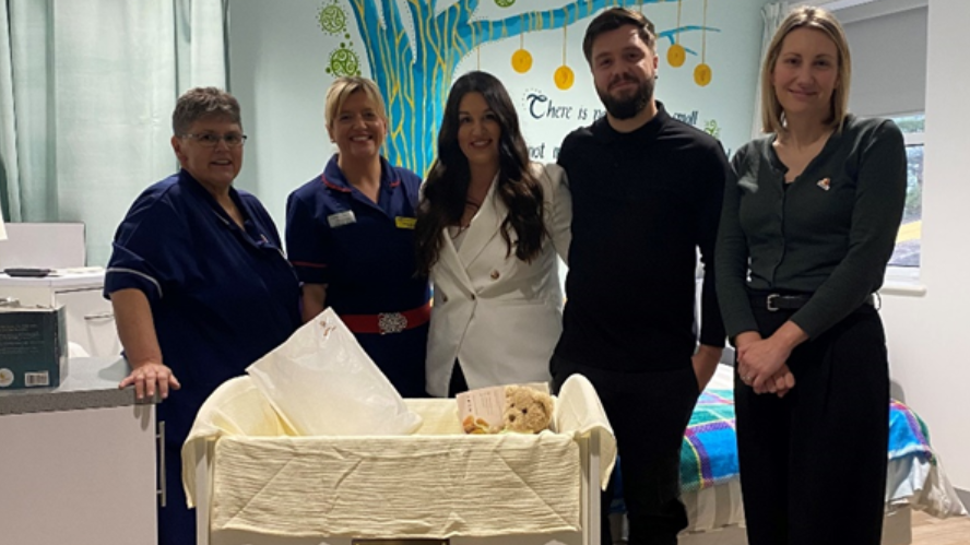 Parents Hannah Spearing and Matt Knight are standing with the staff at the maternity unit at Noble's hospital, and a woman from the Abigail's Footsteps charity. They're standing together in a ward behind a cot covered in blankets with a teddy bear inside. You can see a tree mural behind them on the ward's wall.