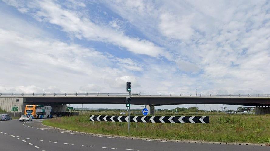 Testo's roundabout. It is a big roundabout with grass. There are traffic lights, signs, two cars a bus.