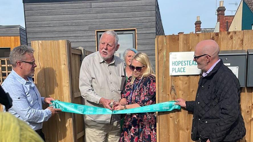 An elderly man wearing a cream-coloured shirt and trousers standing next to a blonde woman with sunglasses and a floral dress. They are cutting a turquoise ribbon in front of Hopestead Place being held by a bald man in a navy jacket and a man with grey hair and a blue shirt. 