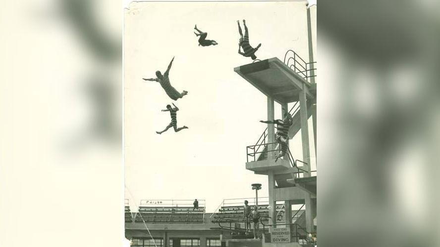 A group of people wearing striped swimming costumes are pictured diving into the pool at the stadium from a high board, in this black-and-white photo.