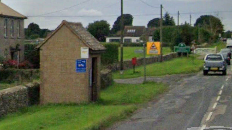 A stone bus shelter on a grassy verge beside a busy road leading into Minchinhampton, with cars passing by on Cirencester Road.
