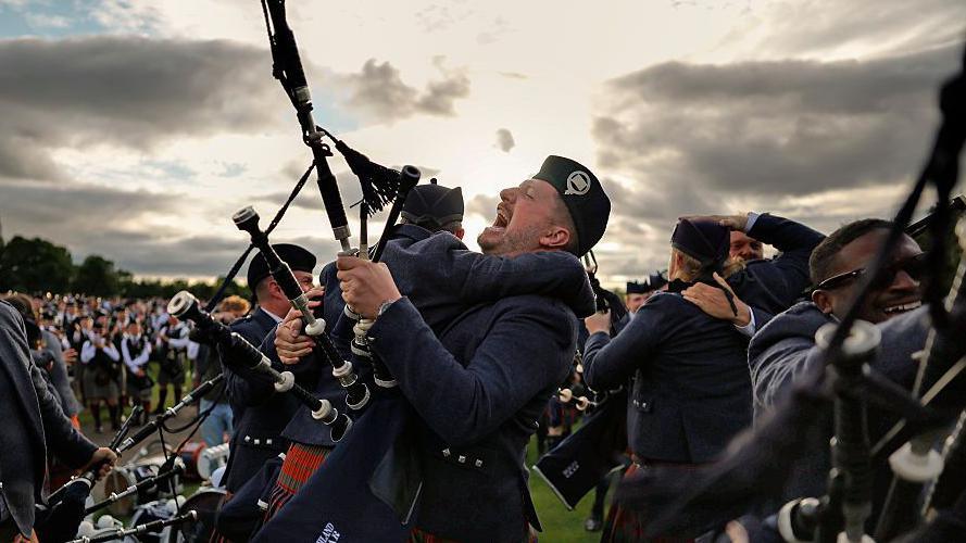 Two pipe band members in matching dark blue uniforms and red-and-green tartan kilts are embracing. One of them is holding bagpipes and his mouth is open in joy. There are other pipers hugging each other around them. There is  low sun behind the clouds creates strong backlighting.