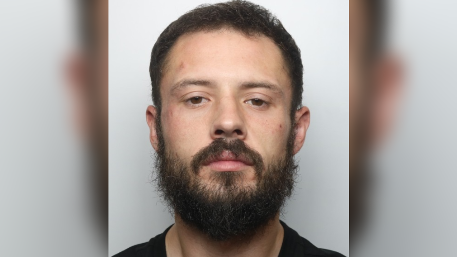 A mugshot of Daniel Kibblewhite. He wears a black t-shirt and has a dark brown beard and hair