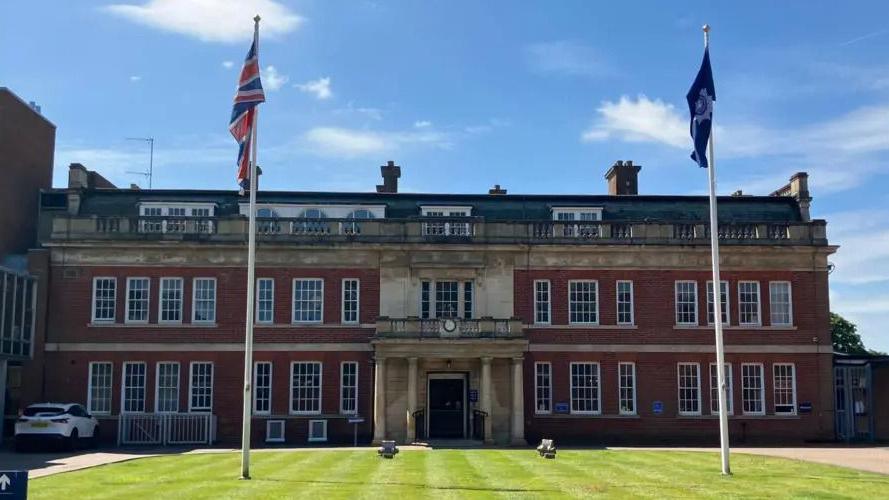 The front facade of Wootton Hall police headquarters. It is a three storey building set in front of a neatly presented lawn with two flag poles. 