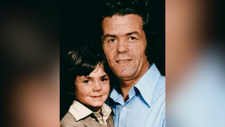 A man and boy smile to the camera. The boy, who sits on the left, wears a brown v-neck jumper with a cream collared shirt. The man wears a blue shirt, with his top button un-done. Both have short dark-brown hair