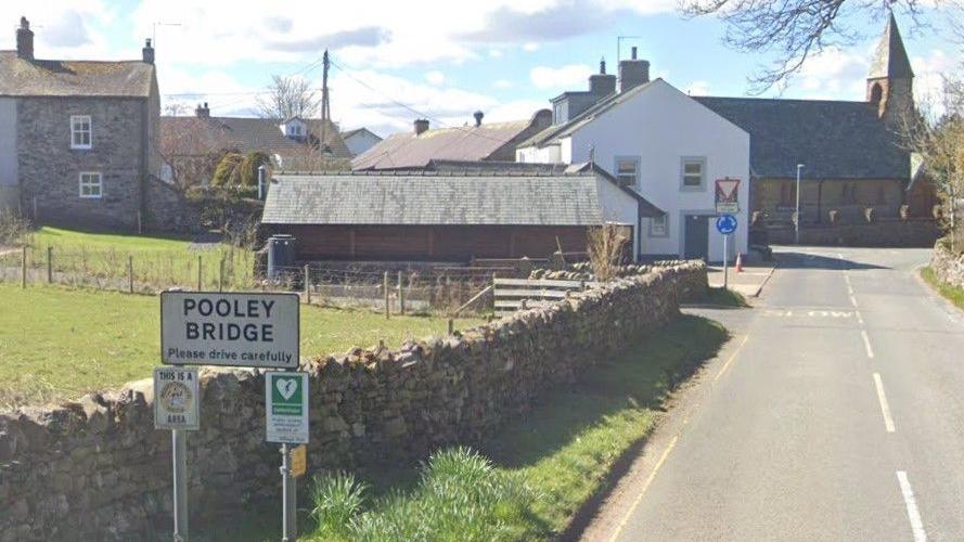 A Pooley Bridge sign at the entrance of the village. There is a small grass field behind  a stone fence with several house further down on the left side of the narrow two lane road. There is a blue roundabout sign near the end of the road and yellow marking reading "slow". 