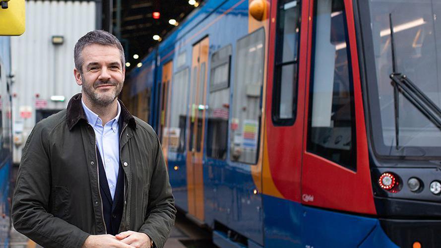 A man with short cropped hair is wearing a white open necked shirt with a coat and is standing by a Supertram