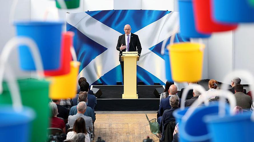 An image from John Swinney's speech - the first minister is behind a yellow lectern, in front of a Saltire backdrop, and there are colourful buckets hanging out of focus in the foreground